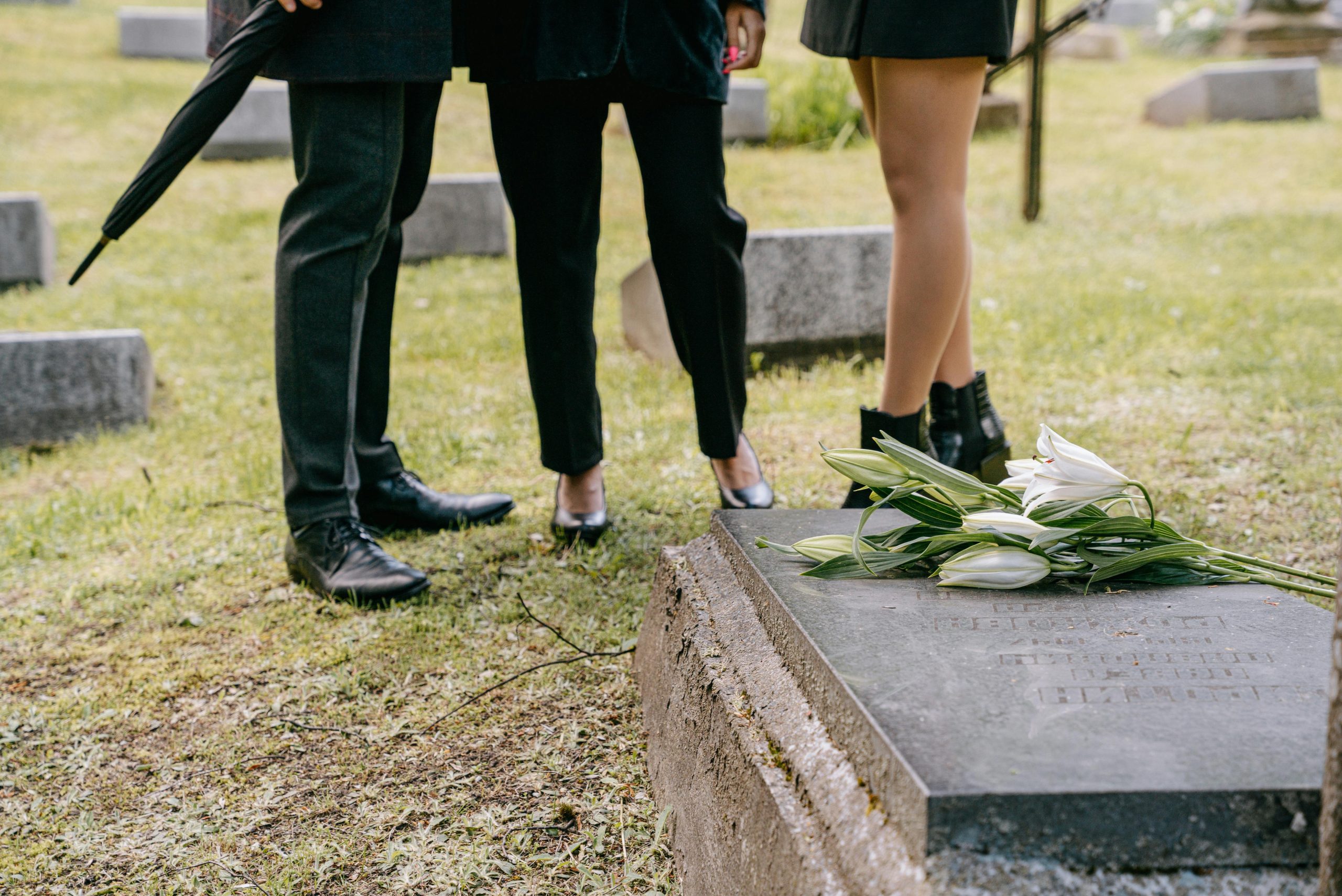 Two individuals in black standing by a grave with white lilies, evoking themes of remembrance.