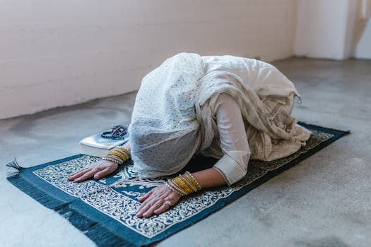 A woman in traditional clothing bowing in prayer on a prayer rug indoors.